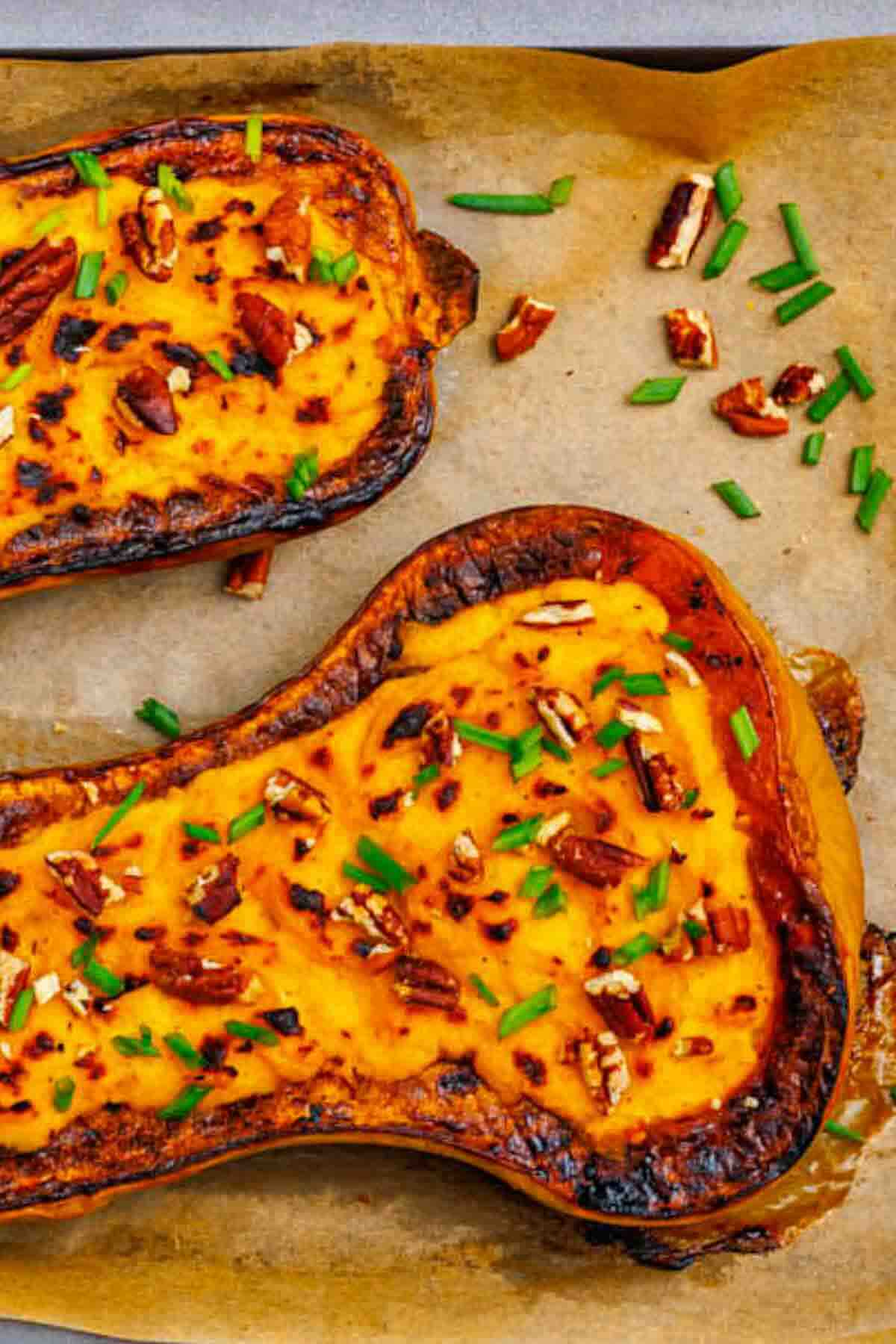 Halved roasted butternut squash on parchment paper, topped with chopped pecans and green herbs.