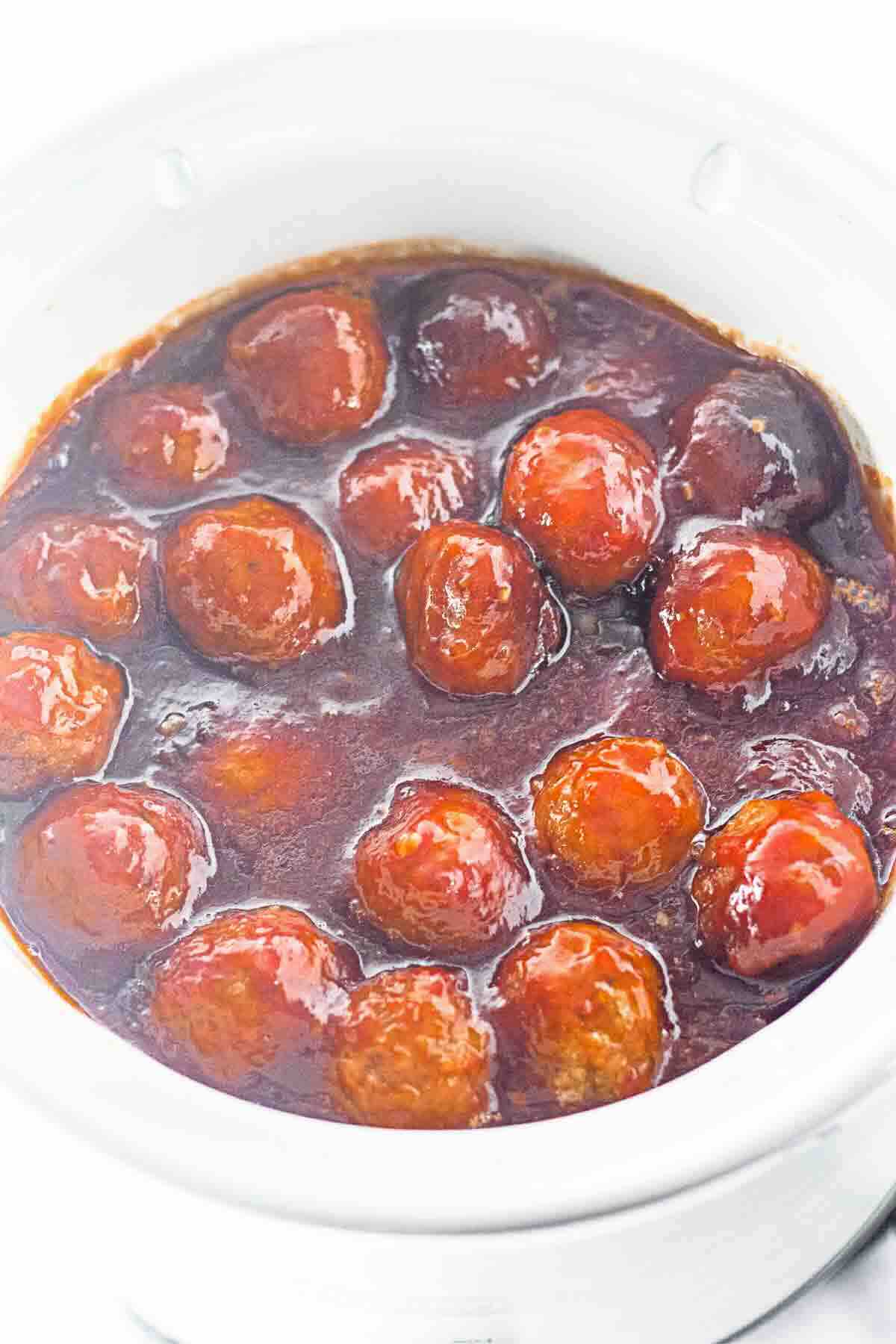 Meatballs coated in a dark brown sauce are shown in a white slow cooker.