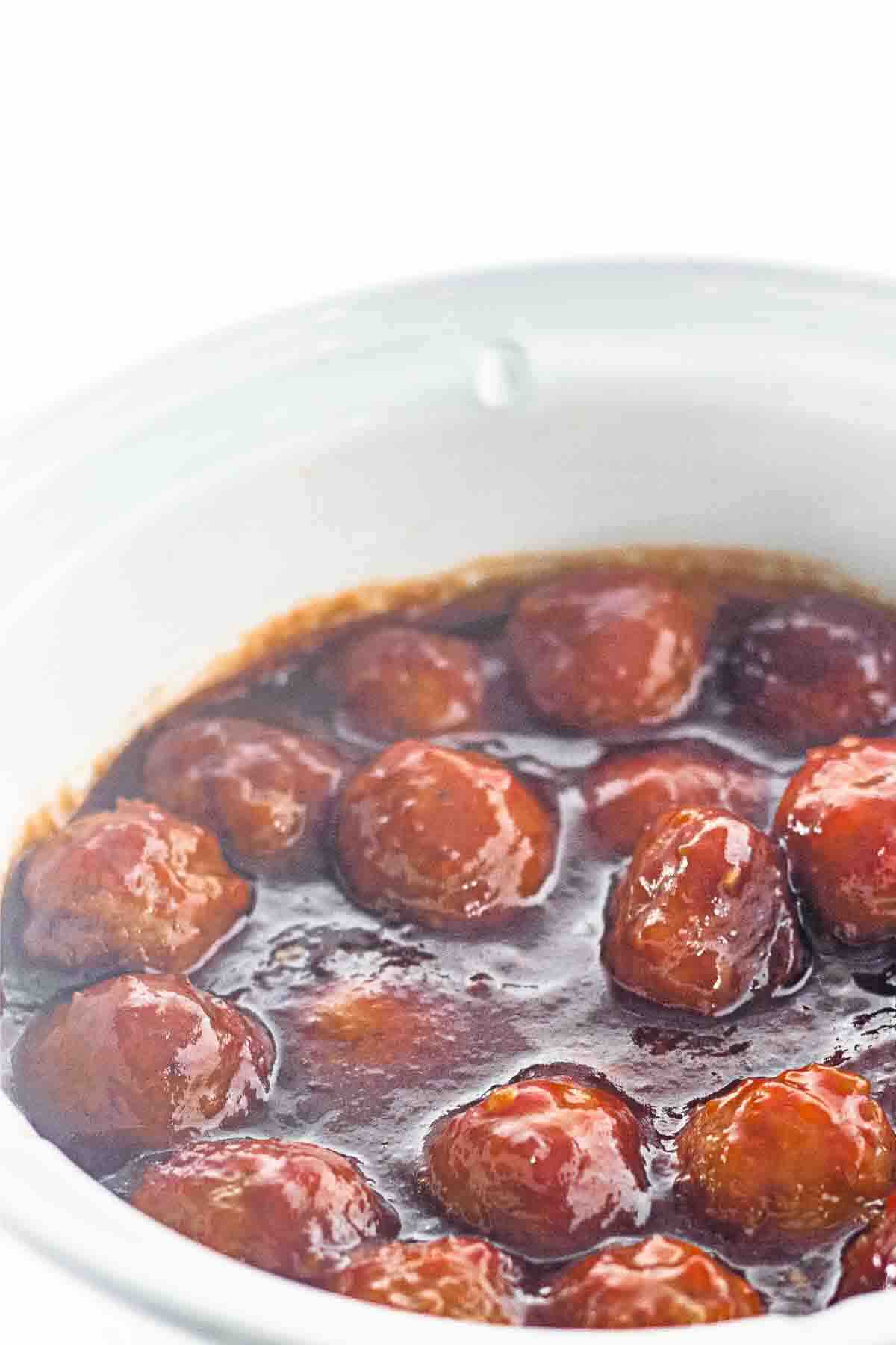 Meatballs coated in a dark sauce simmering inside a white slow cooker.