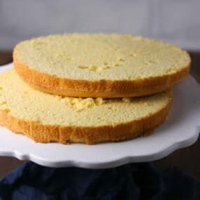 Two round yellow sponge cake layers stacked on a white cake stand, ready for assembling or decorating.