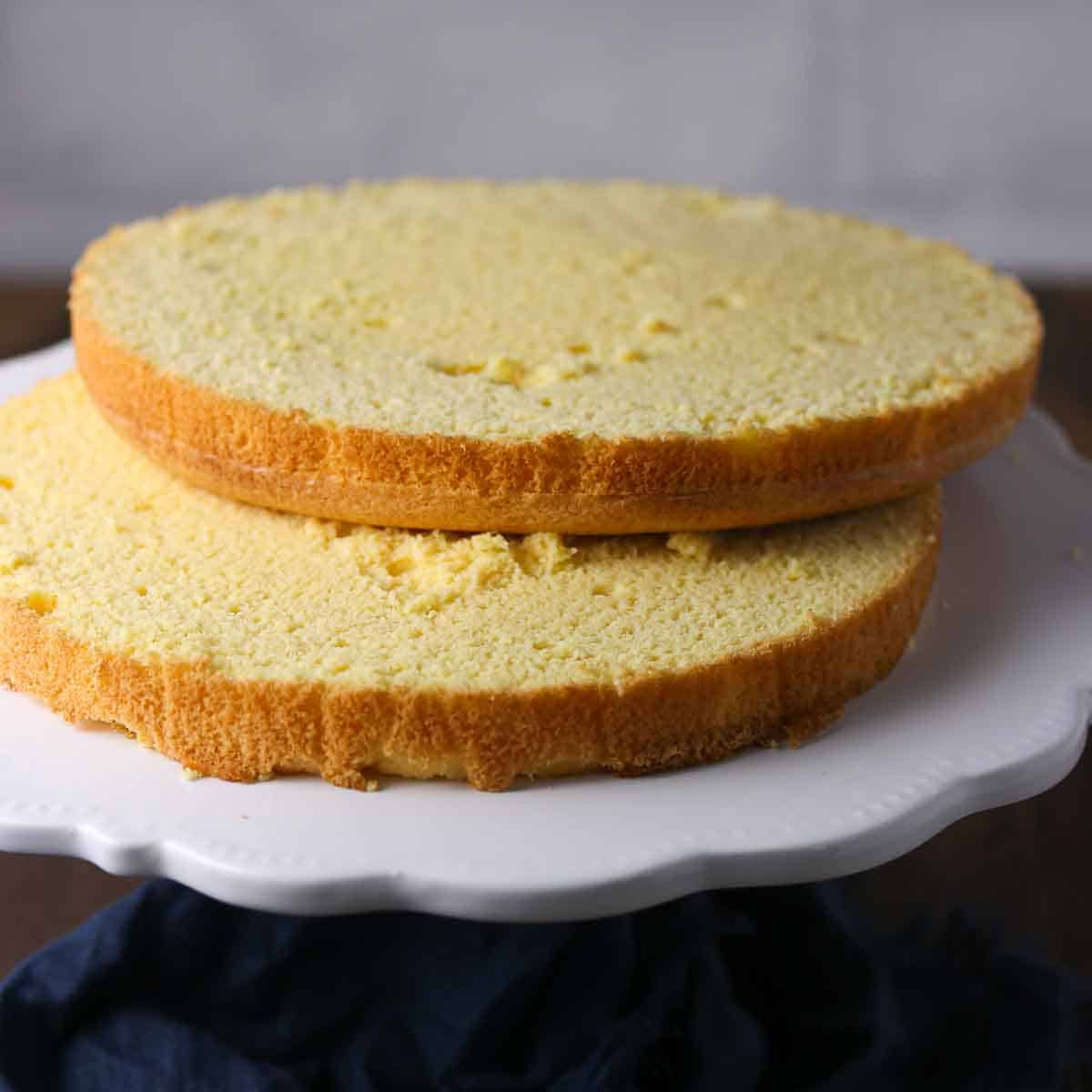 Two round yellow sponge cake layers stacked on a white cake stand, ready for assembling or decorating.