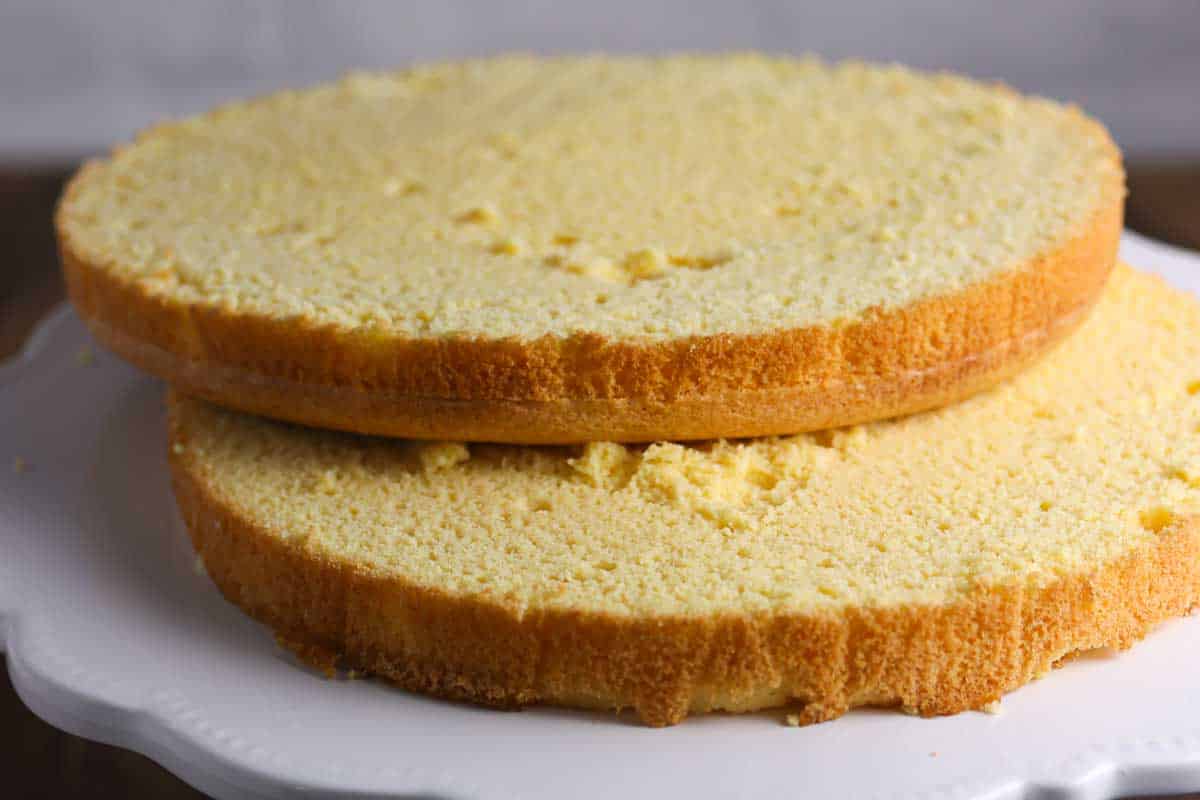 Two plain, round sponge cake layers are stacked on a white cake stand, ready for decorating or assembling into a finished cake.