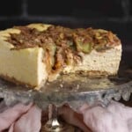 A partially sliced cheesecake with a crumbly topping sits on a metal cake stand, with a light pink cloth underneath.