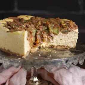 A partially sliced cheesecake with a crumbly topping sits on a metal cake stand, with a light pink cloth underneath.