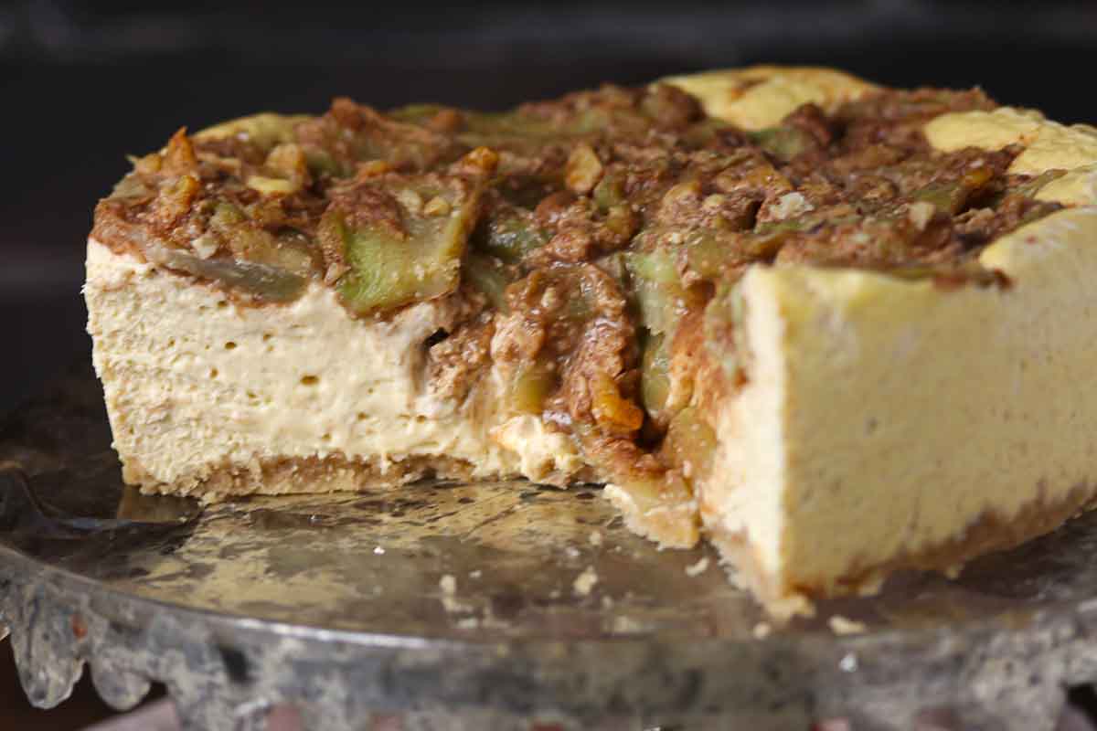 A partially sliced cheesecake topped with apple slices and crumble sits on a glass cake stand.