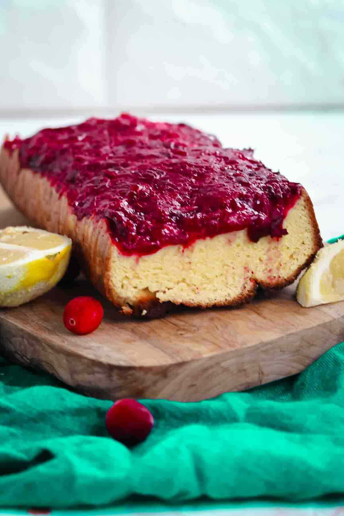 A loaf cake topped with cranberry glaze on a wooden board with lemon wedges and whole cranberries.