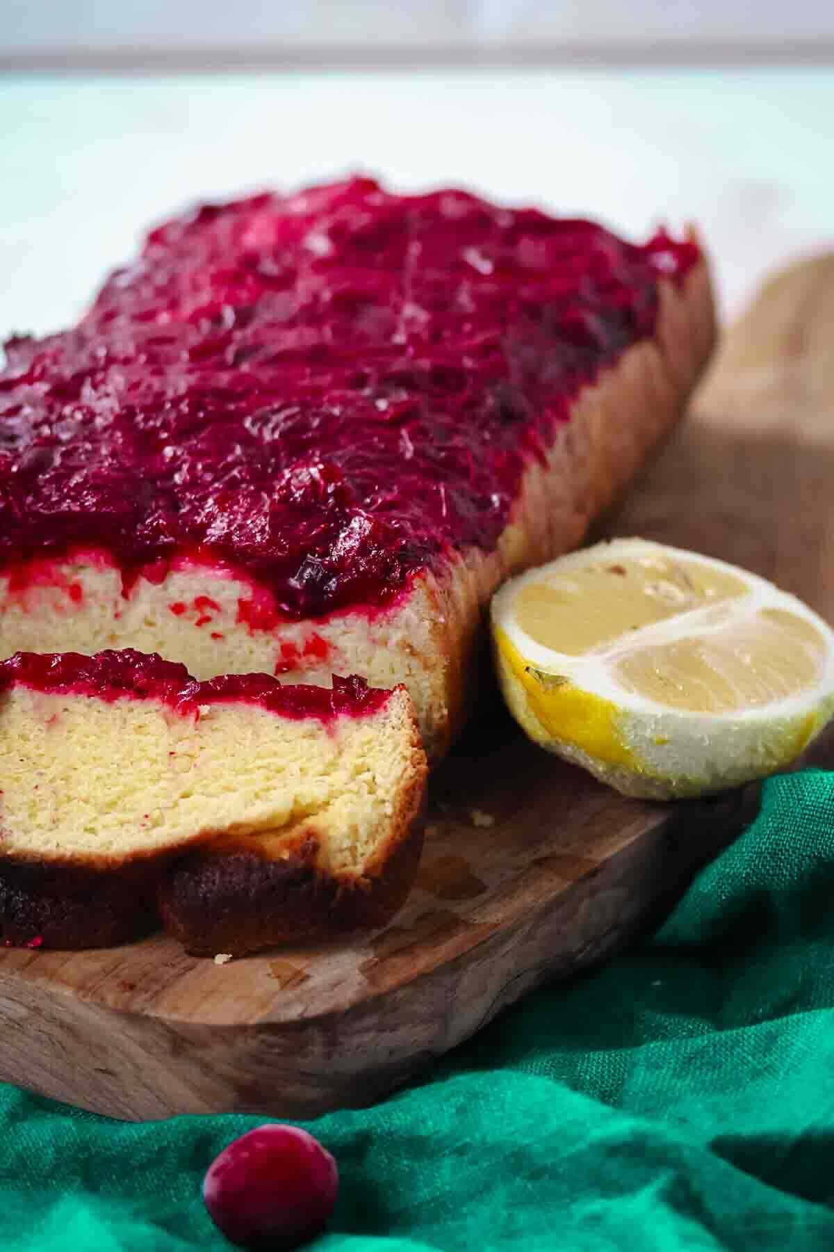 Loaf cake topped with cranberry glaze, with a slice cut and a halved lemon on a wooden board.