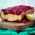 Loaf cake topped with cranberry sauce on a wooden board with fresh cranberries and lemon wedges.