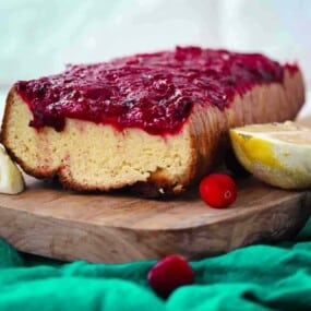 Loaf cake topped with cranberry sauce on a wooden board with fresh cranberries and lemon wedges.